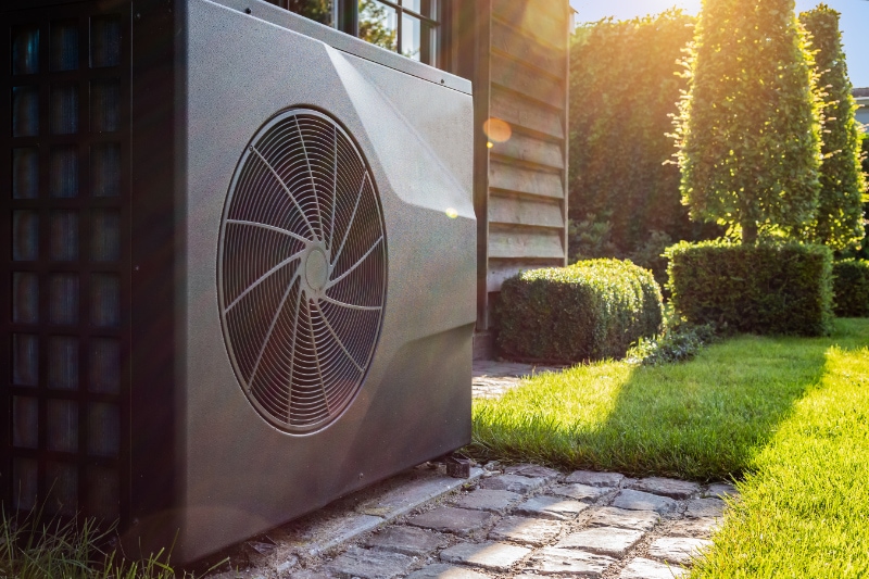 Transitioning Your Heat Pump. Close-up of black full inverter heat pump outside in the garden, near wooden pool house on a sunny day. Lens flare on the image.